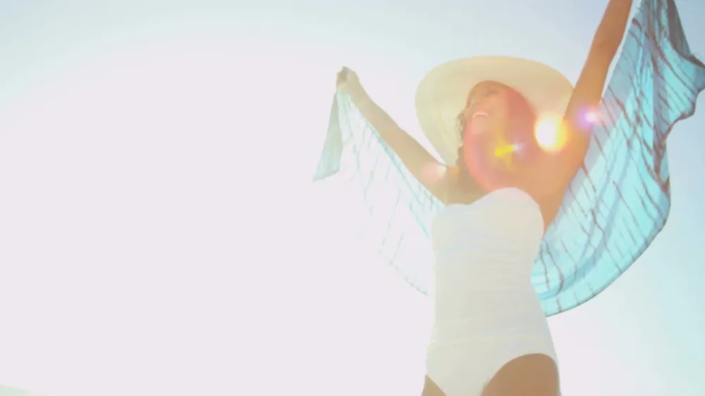A woman in a white swimsuit stands under bright sun, wearing a wide-brim hat and a sheer blue cover, arms raised joyfully.