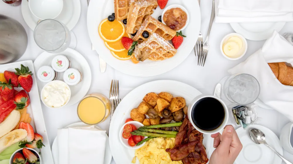 A breakfast spread with waffles, croissants, eggs, bacon, pancakes, fruit, pastries, coffee, orange juice, and a colorful plate of sides.