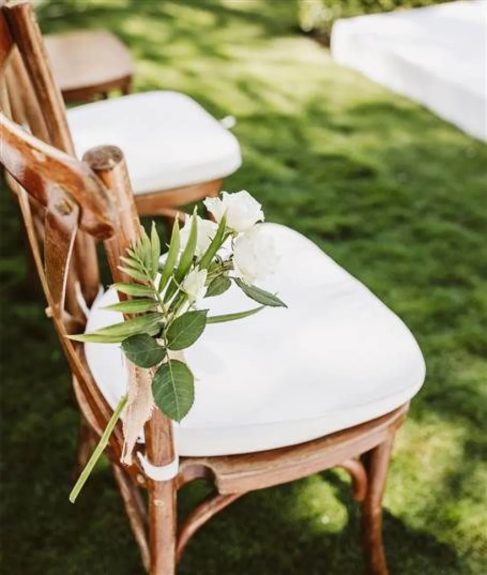 A wooden chair with a white cushioned seat outdoors, decorated with a small green leafy sprig tied to the chair back using twine.