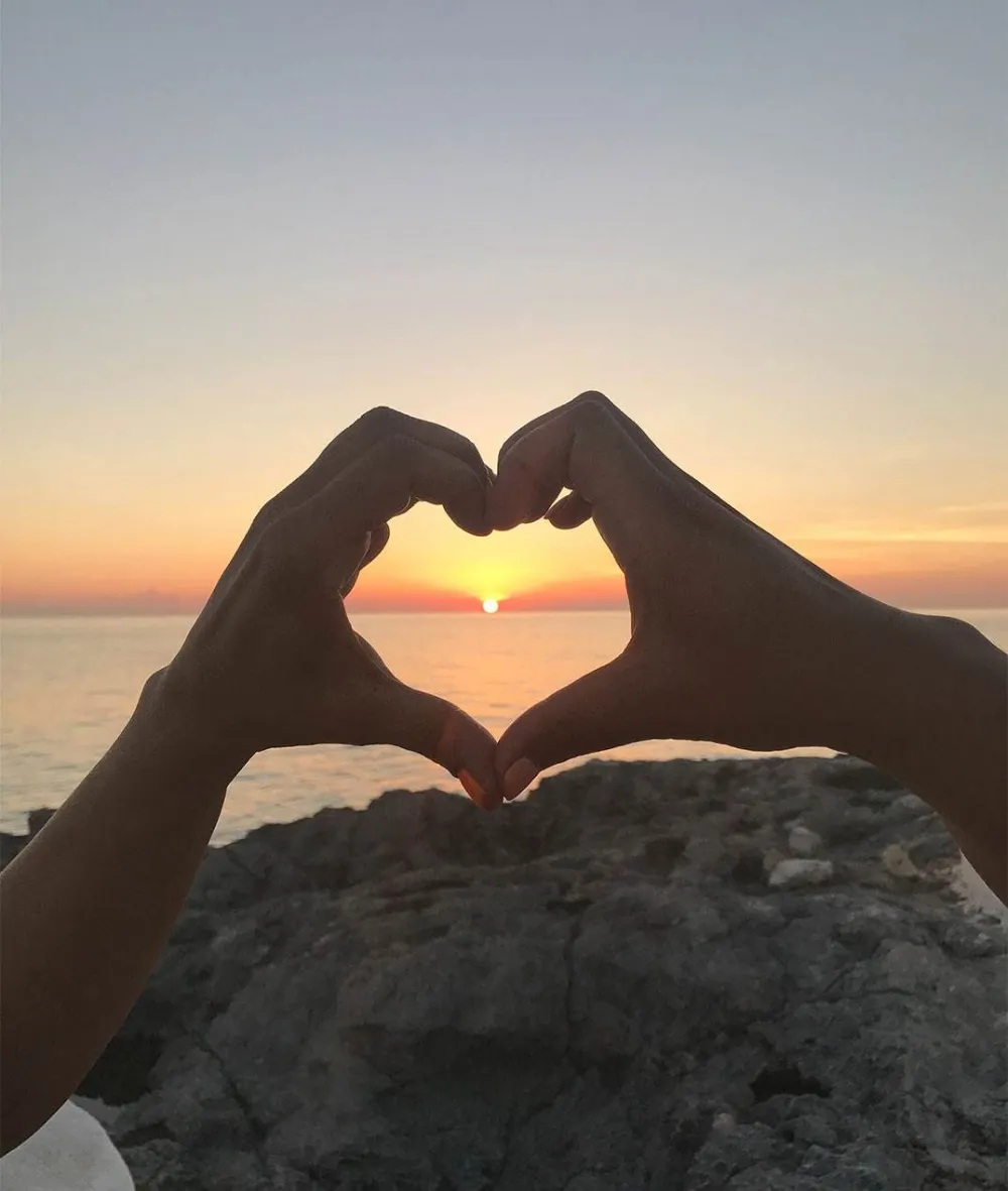 Two hands form a heart shape around a sunset over the ocean, with a rocky shore in the foreground, capturing a romantic seaside moment.