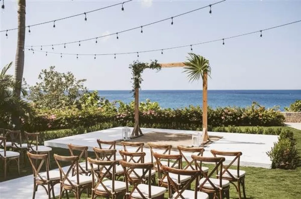 Outdoor beach ceremony setup with wooden chairs, string lights, floral arch, and ocean view in the background, ready for guests.