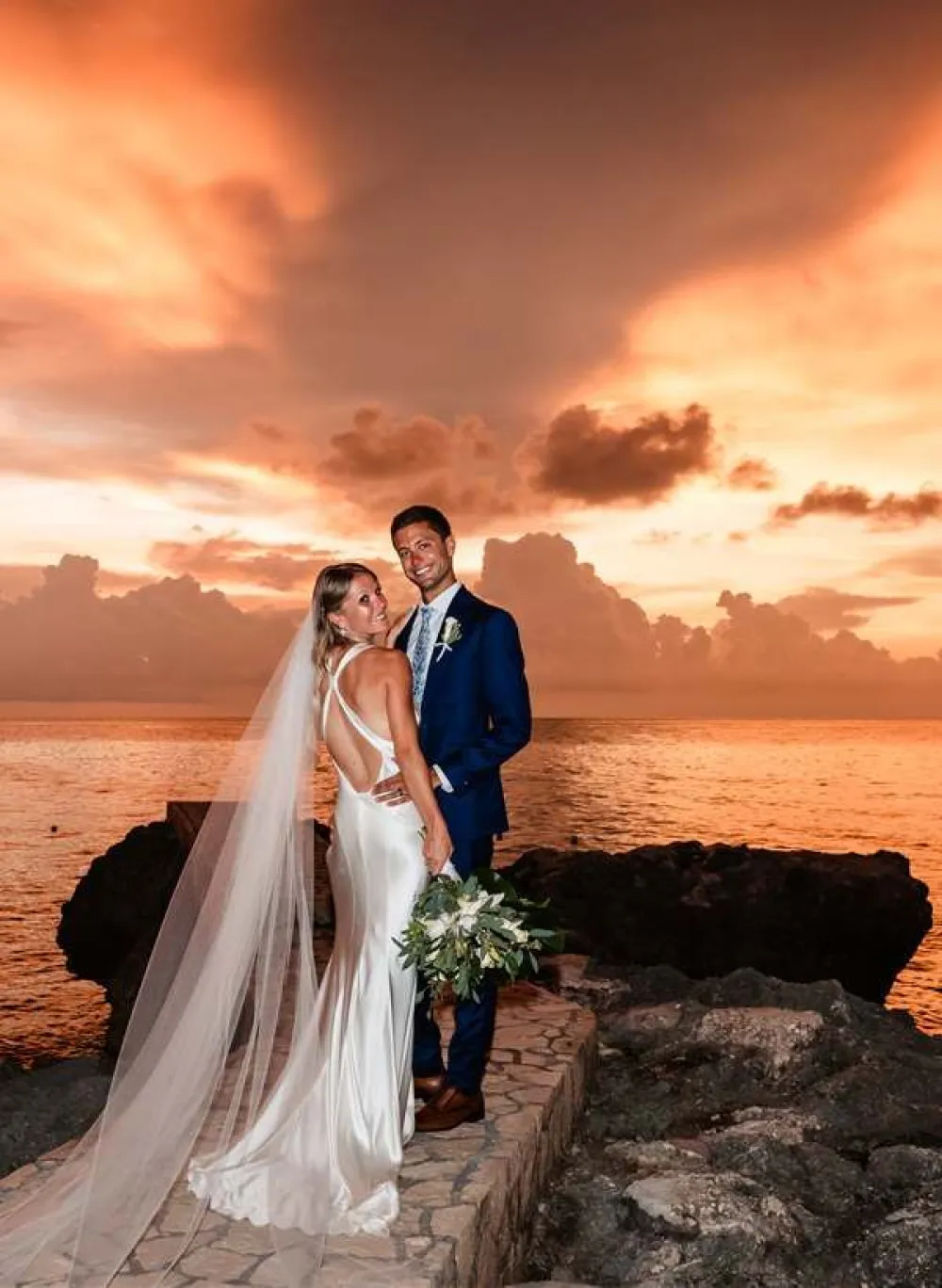 A bride and groom stand on rocks by the sea at sunset, the bride in a long white gown with veil, holding greenery bouquet, smiling at camera.