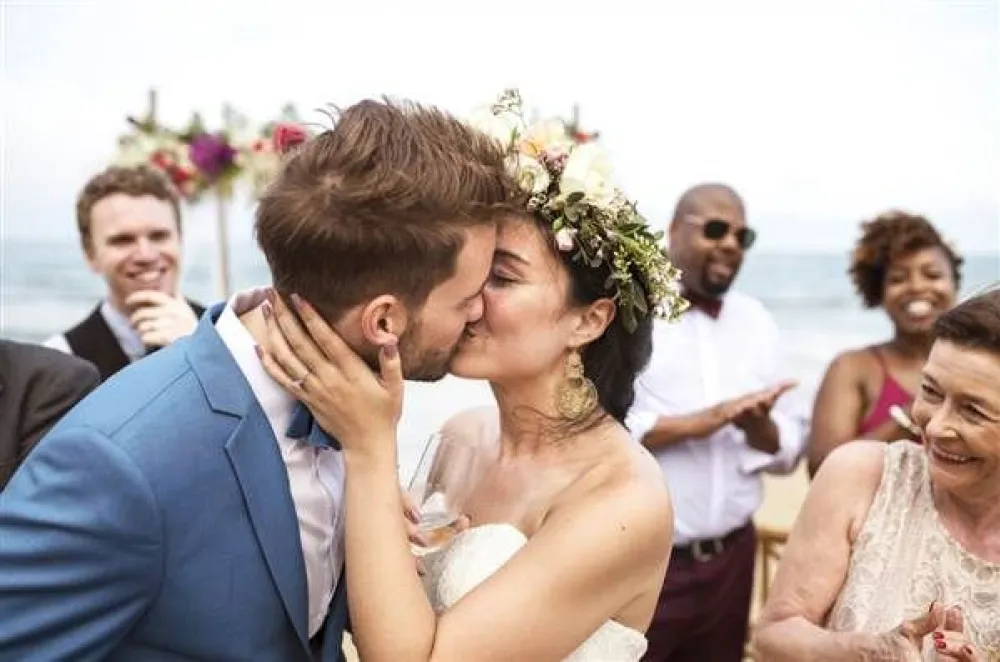 A bride and groom kiss at their outdoor wedding ceremony, guests celebrate in the background by the sea.