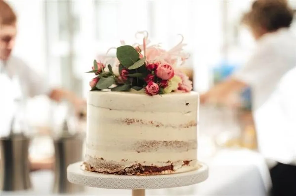 A simple naked cake with light frosting and a floral arrangement topper on a cake stand, set in a bright kitchen.