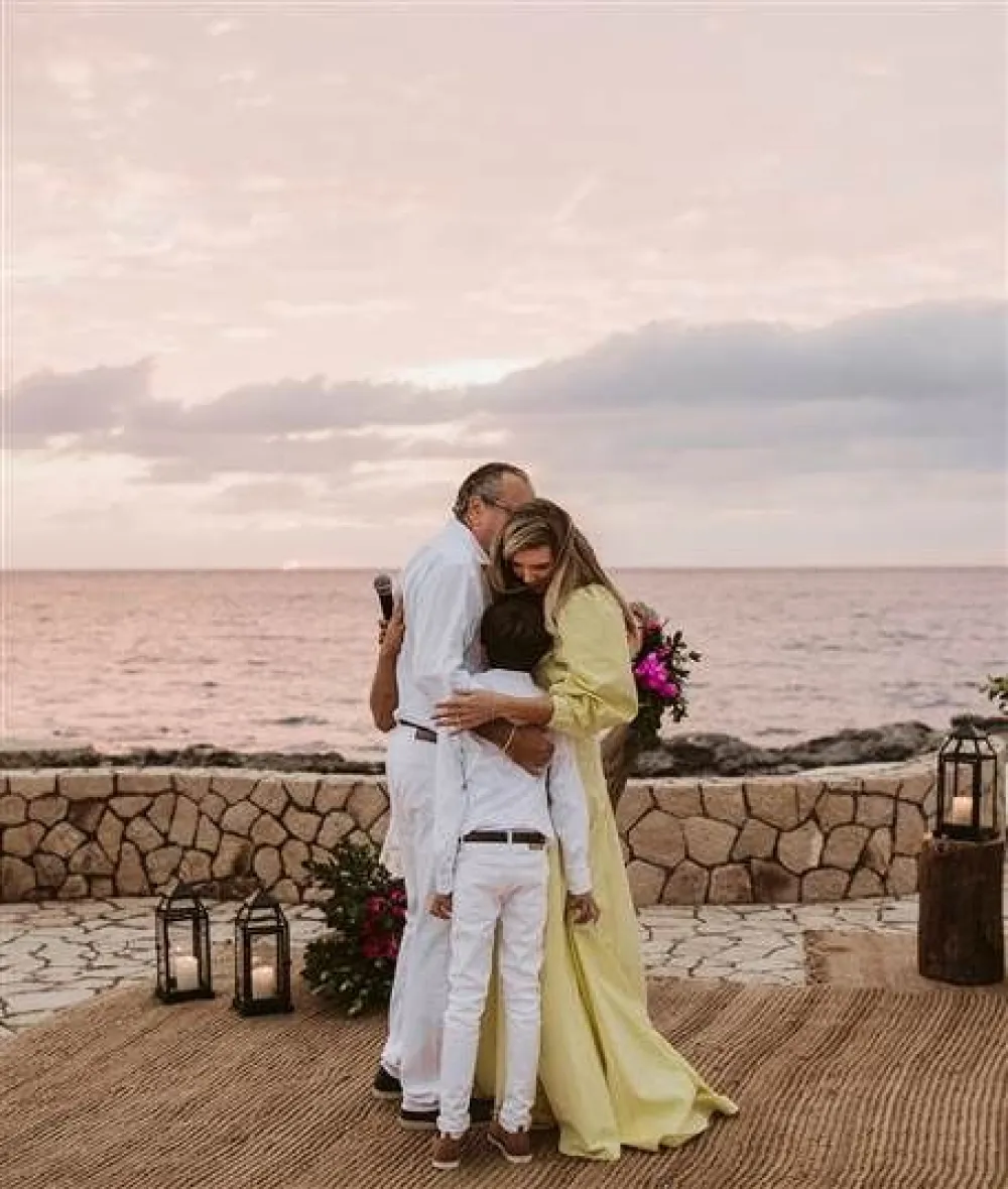 A couple hugs on a seaside patio at sunset, sharing a tender moment with lanterns and a stone barrier in the background.