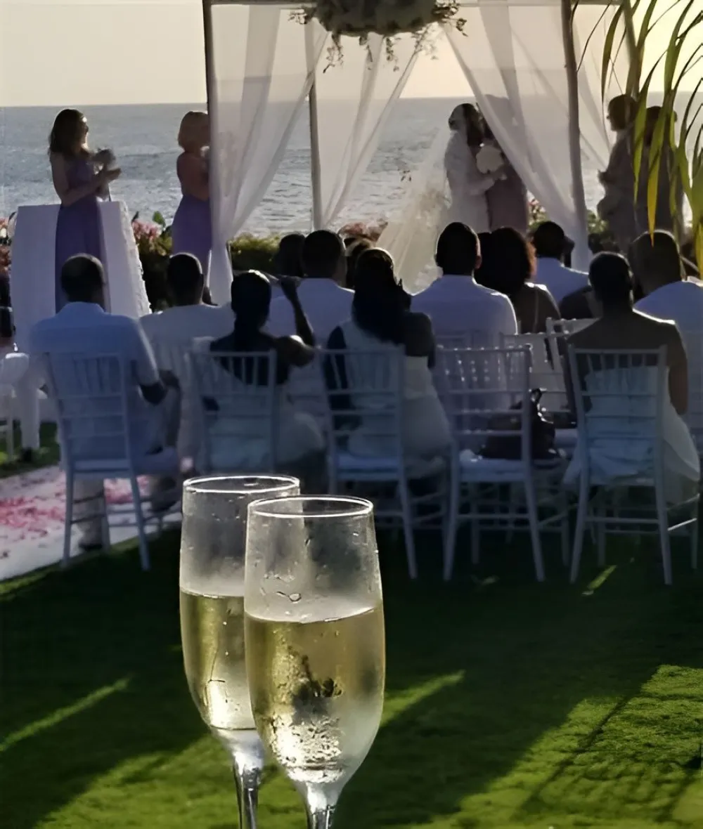 A wedding ceremony outdoors on the beach with a white aisle, seated guests, and two champagne glasses in the foreground.