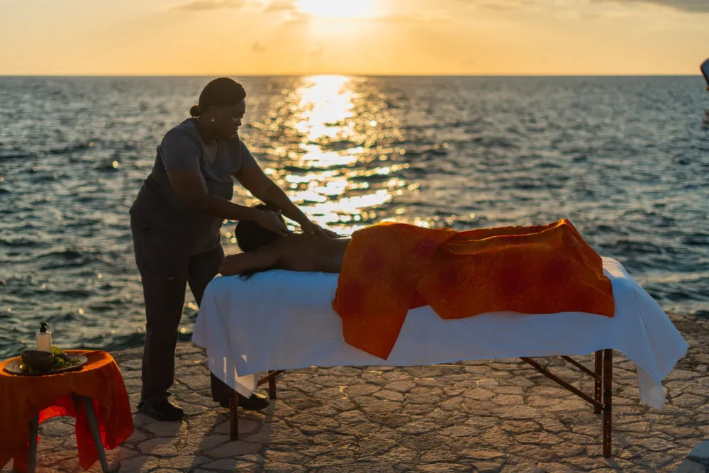 A person giving a massage on a beach at sunset, using a massage table with a white sheet and an orange towel, by the ocean. Top it at 140 characters, always ending the sentence.