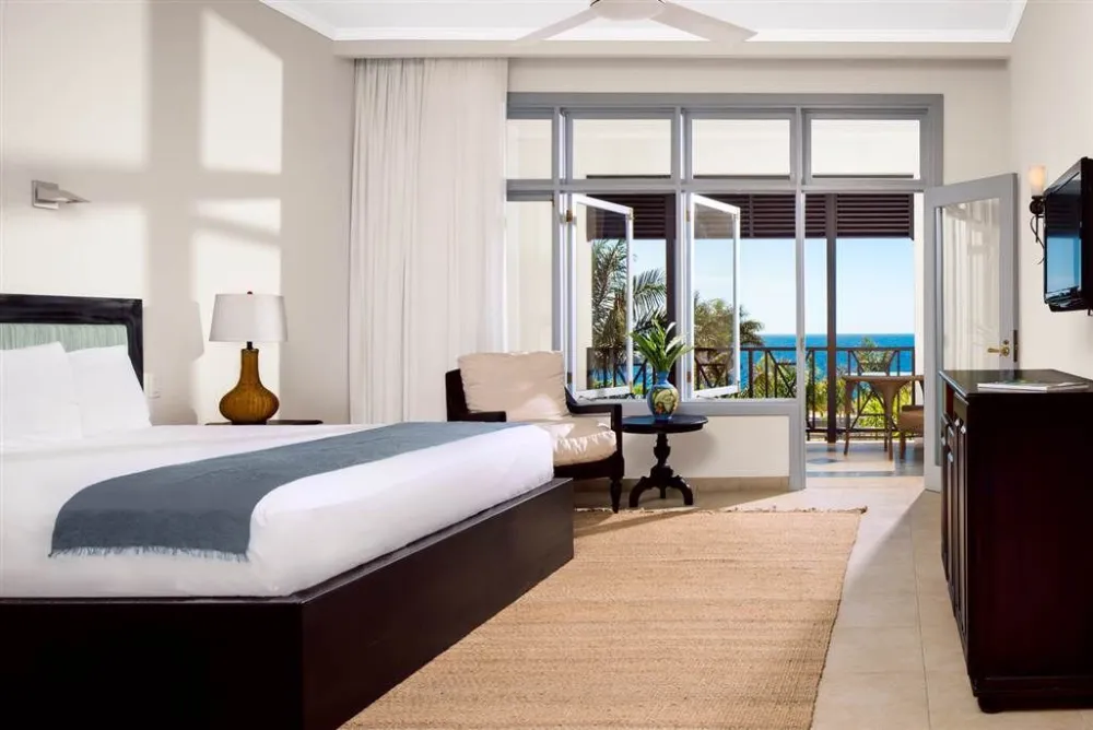 A bright hotel room with a large bed, nightstand, chair, and a view of a balcony overlooking the ocean and palm trees.