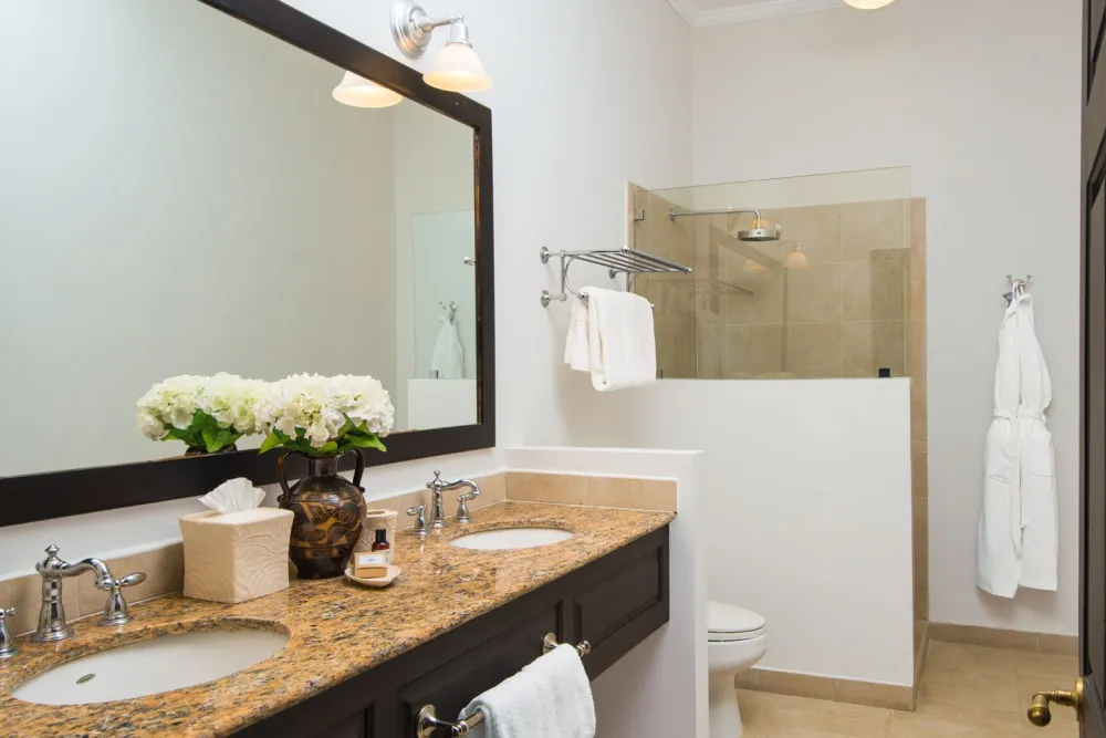 Amodern bathroom with a double vanity, granite countertops, large mirror, and a glass-enclosed shower; towels and flowers add a decorative touch.