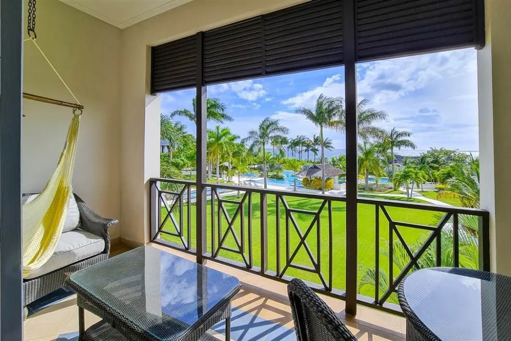 A balcony dining area with wicker furniture overlooks a sunny lawn and a pool-like view framed by palm trees.