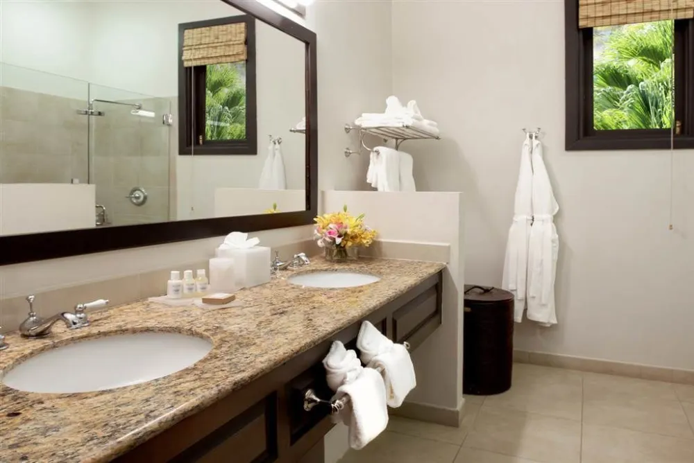A modern hotel bathroom with double granite sinks, toiletries, folded towels, and a glass-enclosed shower.