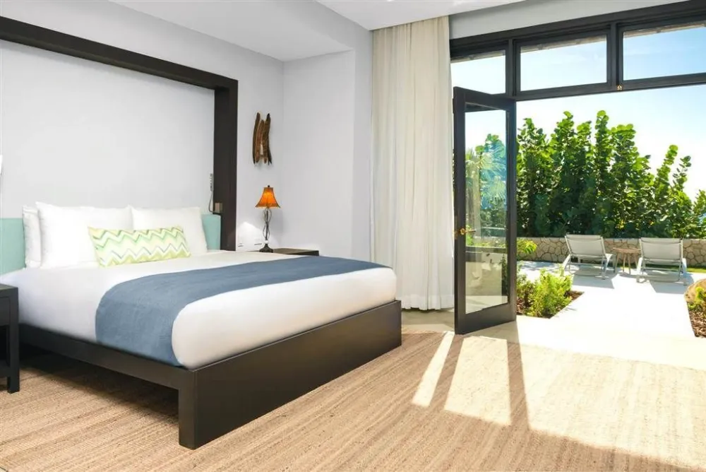 A modern hotel room with a dark wood bed, white bedding, mint-green accents, and a sliding glass door opening to a sunny patio and pool area, ending here.