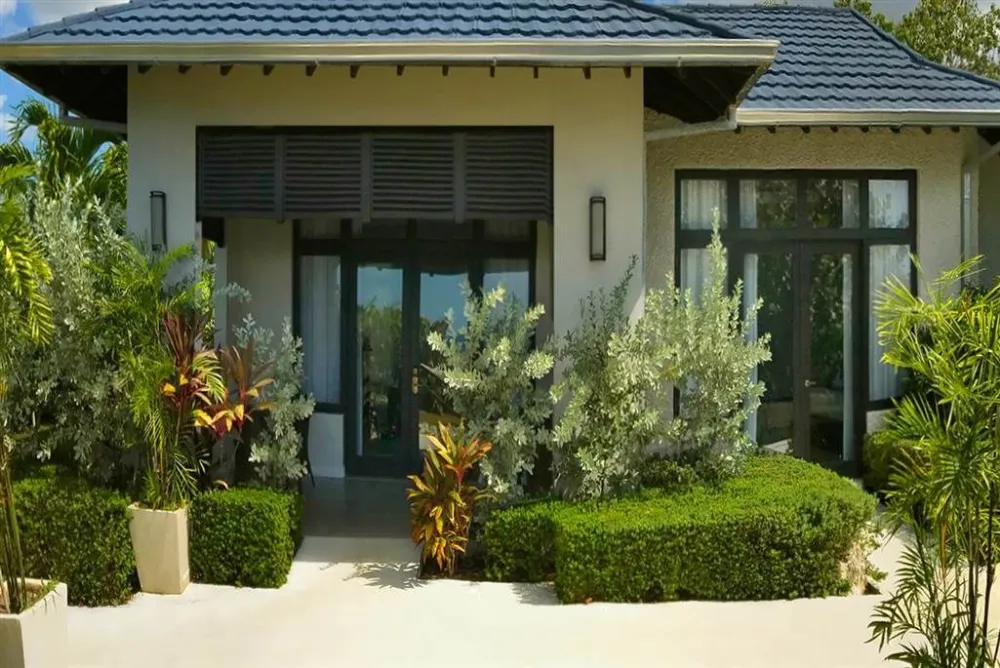 A modern single-story house with large glass doors, tropical landscaping, tidy hedges, and potted plants lining a sunny entrance.