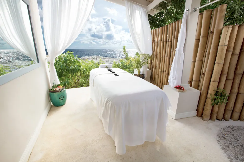A serene outdoor spa setup with a white massage table under flowing curtains, overlooking a distant city view and lush greenery.