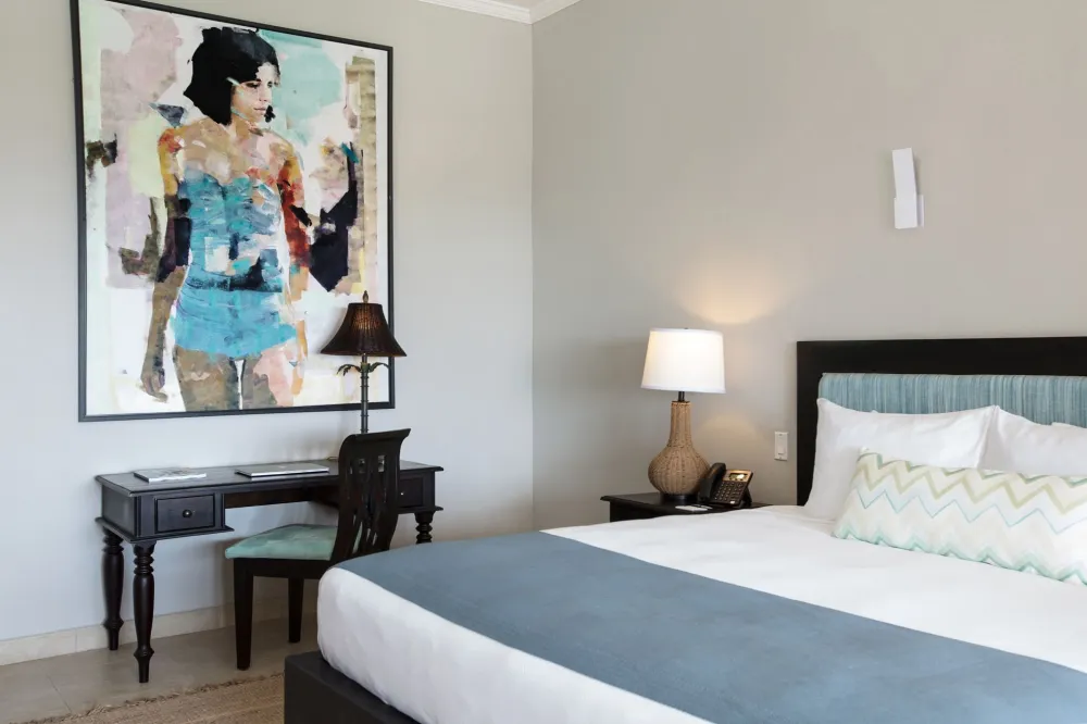 A modern hotel room with a queen bed, gray walls, bedside lamps, a framed abstract painting, and a desk with a chair by the wall.
