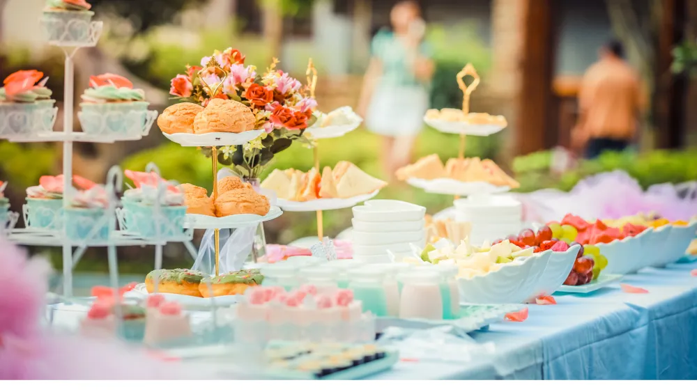 An outdoor garden party buffet with a tiered stand of buns, colorful flowers, and a table of sweets under soft light.