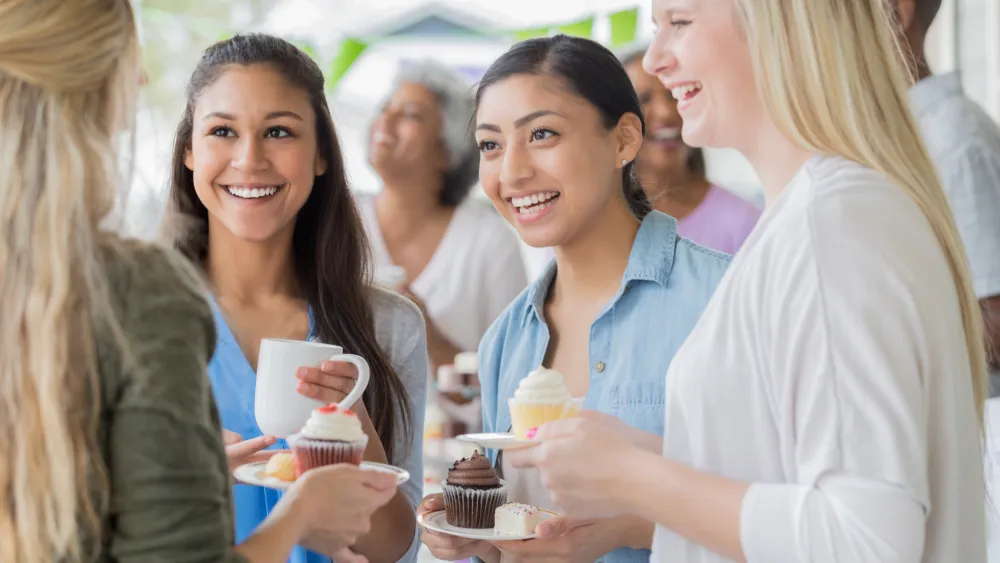 People socializing at a casual gathering, sharing cupcakes and drinks, smiling and chatting in a bright, friendly setting.
