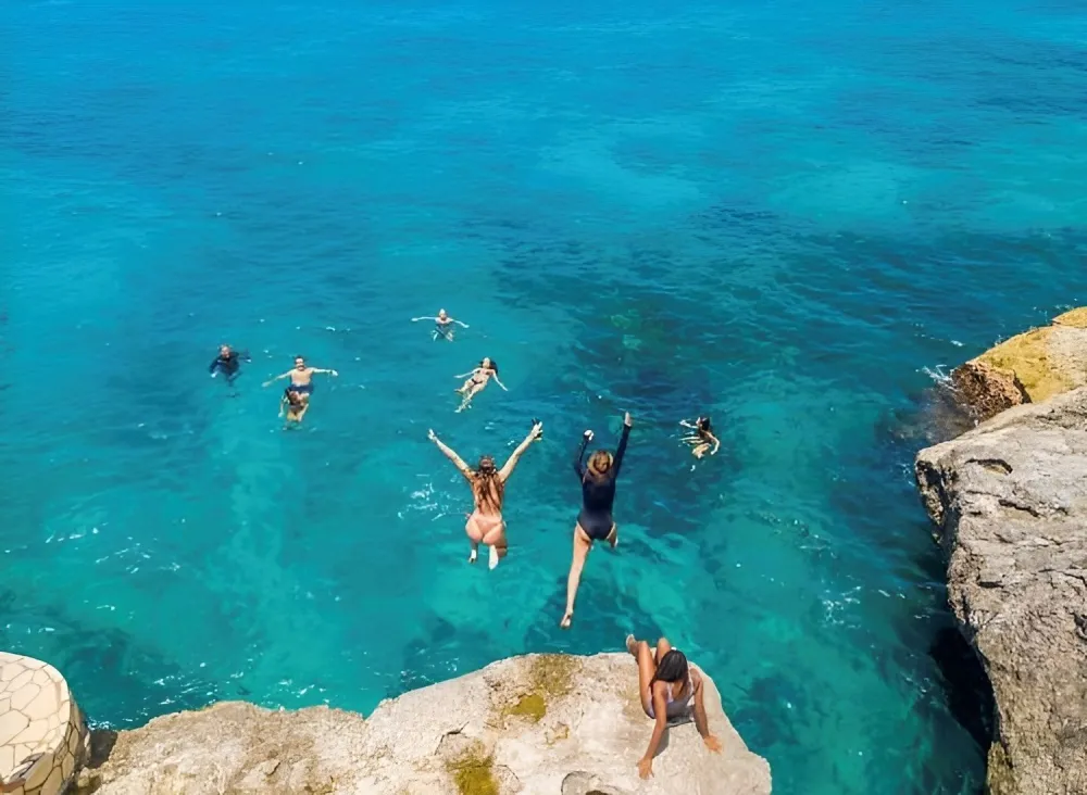 People jump into a clear turquoise ocean from a rocky cliff, swimming and diving in open water.