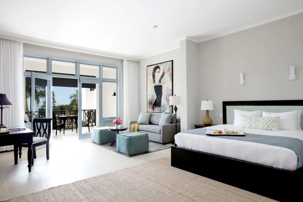 A bright hotel-style suite with a king bed, a seating area, and sliding glass doors opening to a balcony, modern decor, and light neutral tones.
