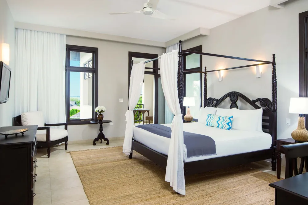 A luxurious tropical hotel room with a dark four-poster bed, white curtains, blue and white bedding, a rug, desk, chairs, and large windows with sheer curtains.