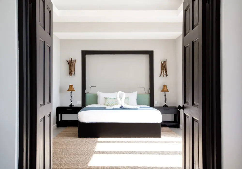 A bright, modern bedroom seen through dark double doors: a low bed with white and green bedding, two matching nightstands, and soft natural light.