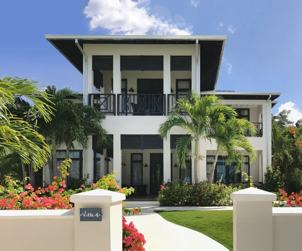 A modern two-story white villa with dark trim, lush tropical landscaping, palm trees, and a curved walkway leading to the entrance.