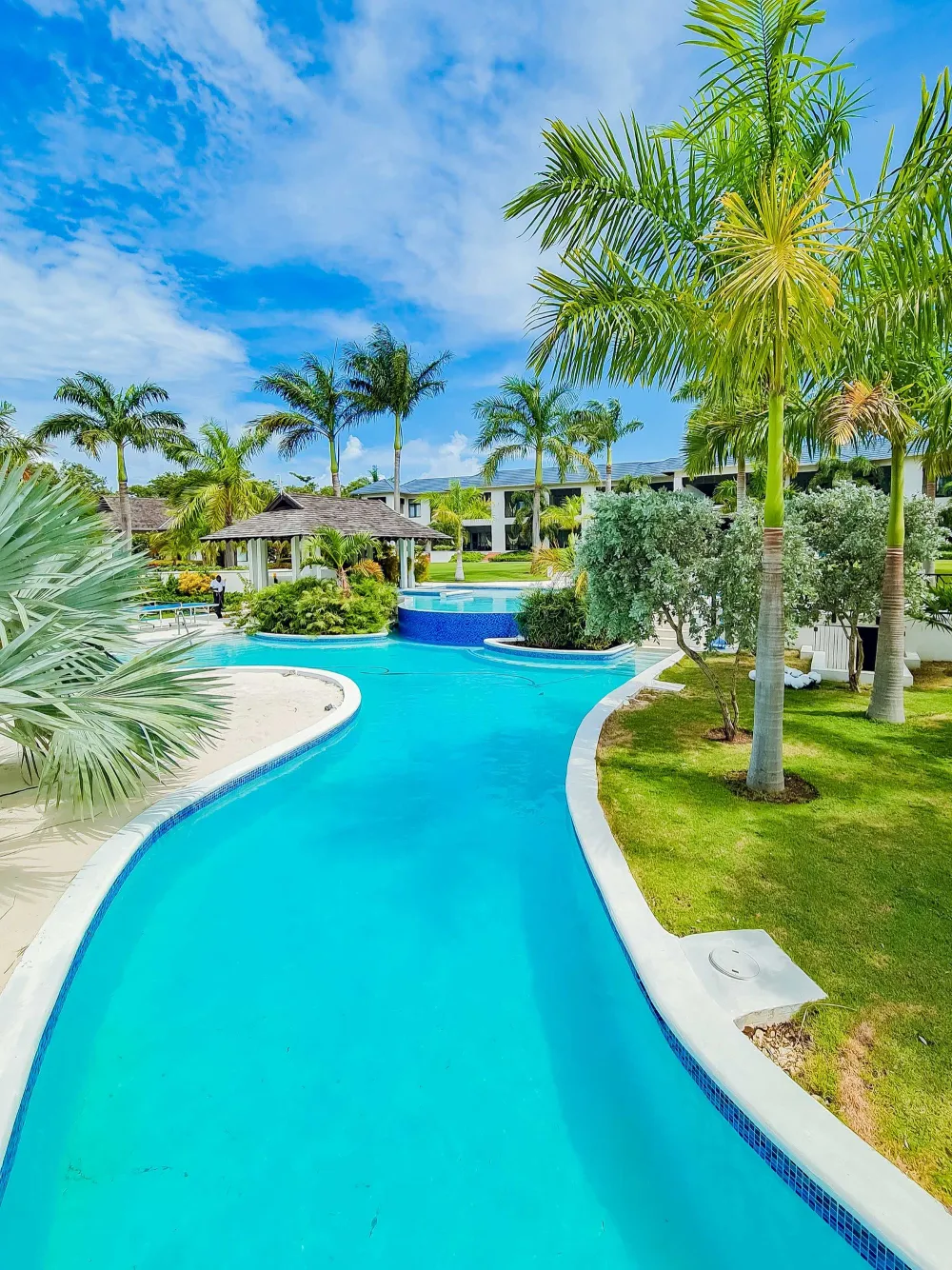 A tropical resort pool winds through palm trees toward a bright blue sky, with a curved slide and turquoise water, inviting relaxation.
