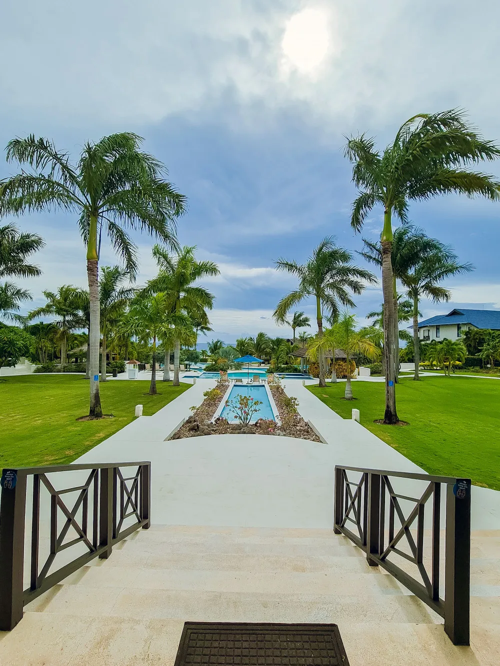 A tropical resort scene with a stone path leading to a pool, flanked by palm trees under a sunny sky, inviting relaxation and sunshine.