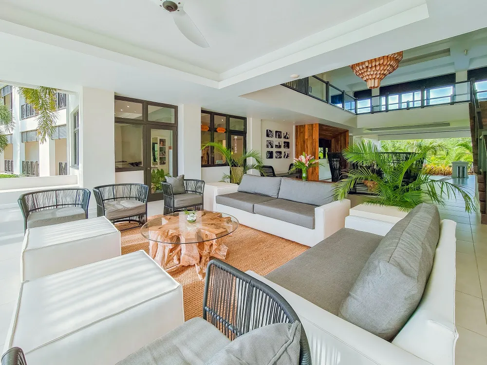 Bright modern patio living room with white sofas, a glass coffee table, ceiling fan, and lush potted plants under skylight.