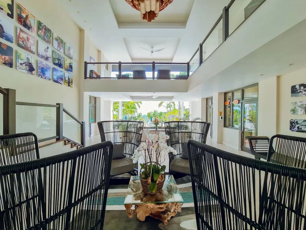 A bright indoor lounge/balcony with black wicker chairs, an orchid centerpiece, a photo wall, and doors opening to greenery.