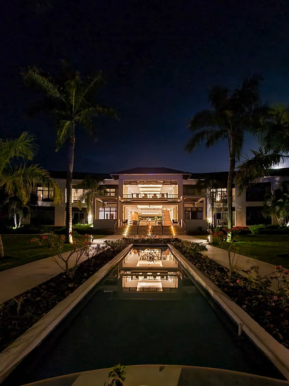 A nighttime scene of a modern house with a bright entrance, palm trees, and a rectangular reflecting pool leading to the lit doorway.