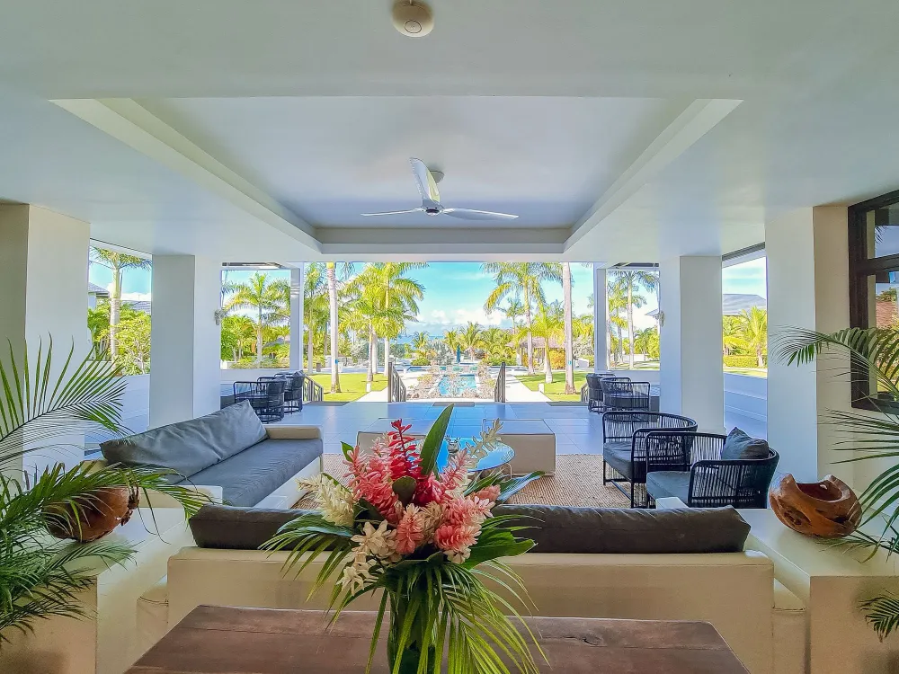 A bright, open living area with white walls, ceiling fan, and large windows showing palm trees and a pool; modern seating and tropical centerpiece.