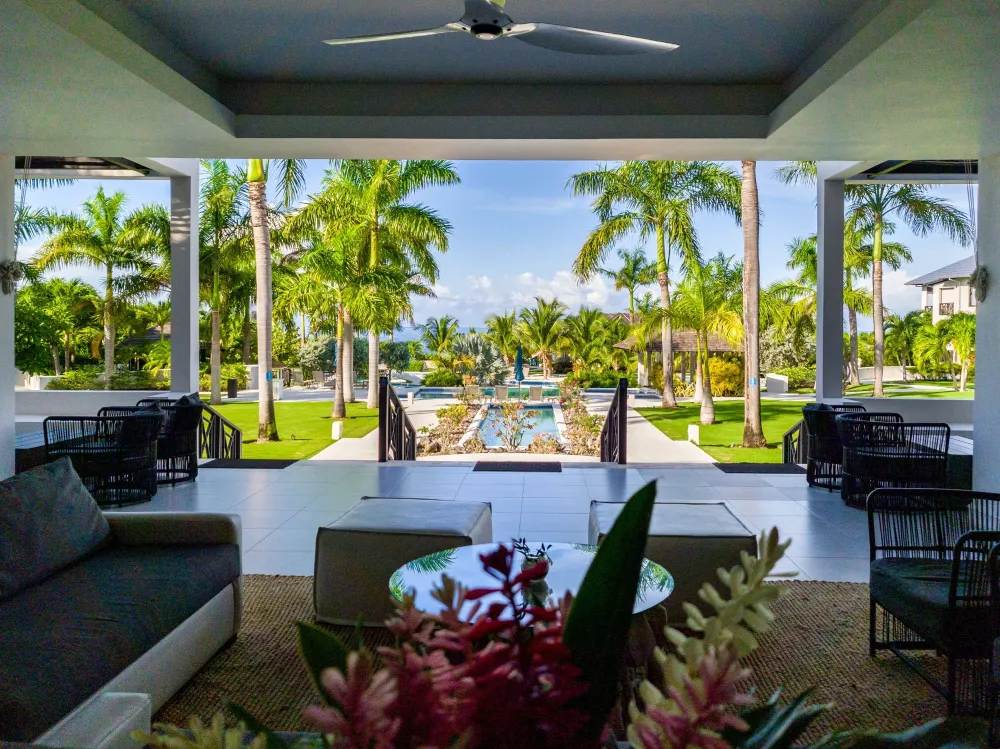 A tropical resort patio with comfy seating, a small pool, lush palm trees, and manicured lawns under a sunny sky.