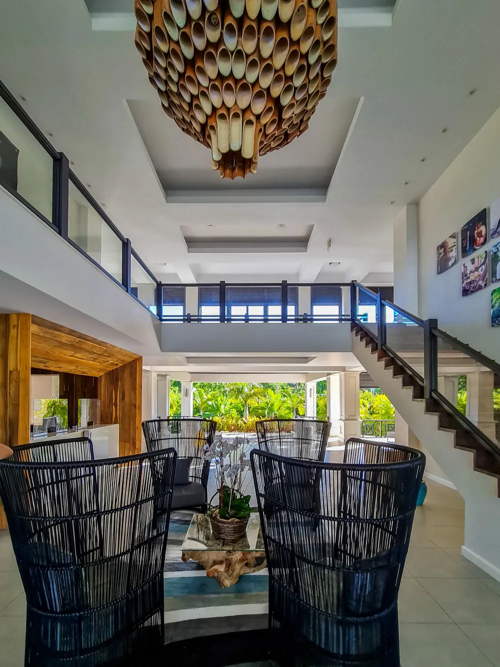 A bright, modern living/dining area with high ceilings, a woven pendant light, glass railing balcony, and stylish black chairs facing a coffee table.