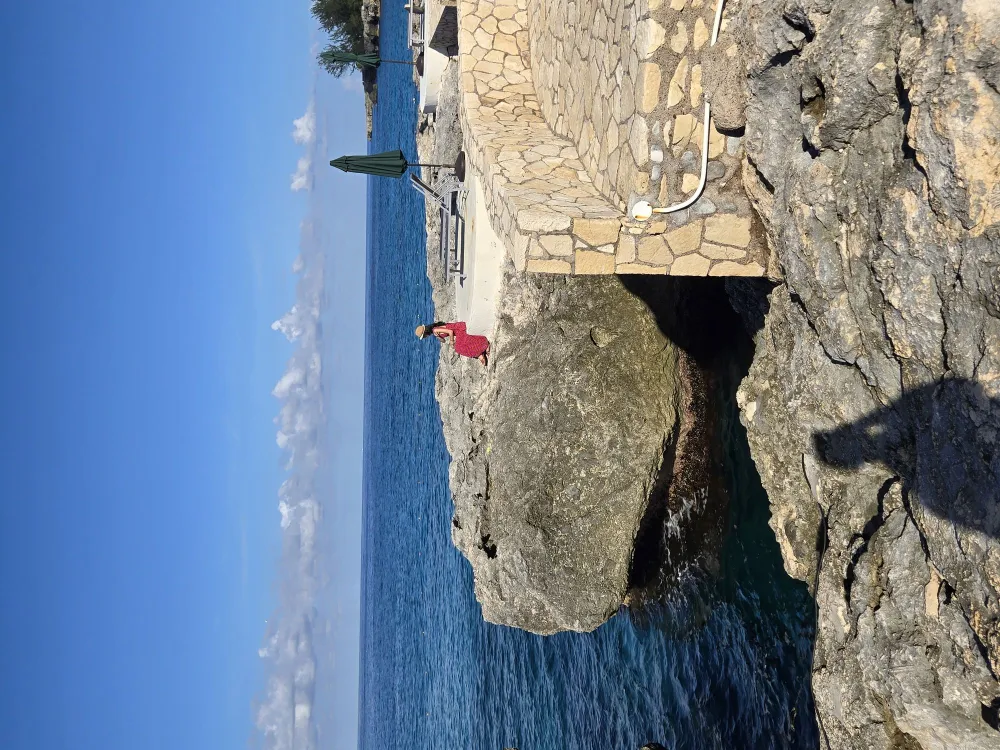 A person in a red outfit sits on a rocky ledge by the sea, with a stone walkway and clear blue water under a bright sky.