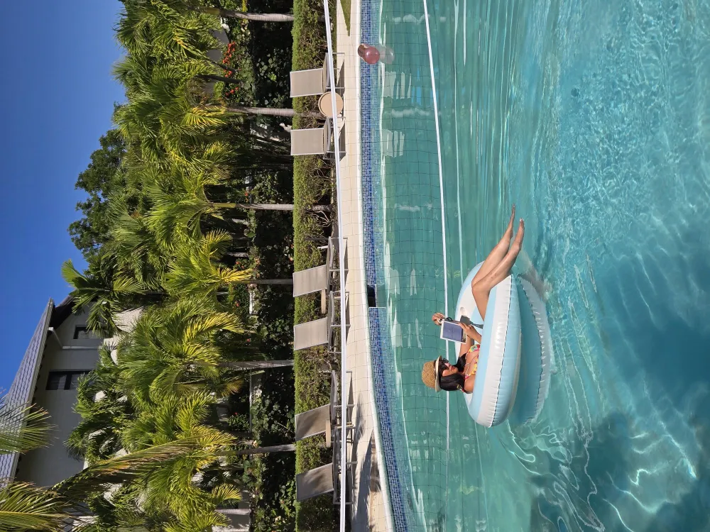 Two people on an inflatable raft in a bright pool, surrounded by palm trees and a sunny resort scene.