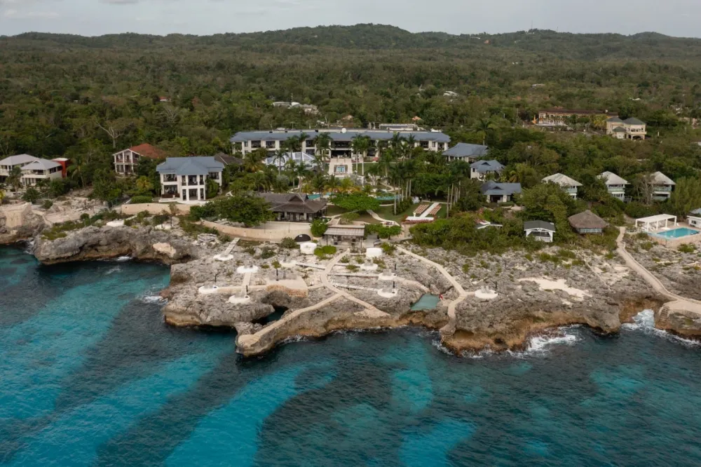A beachfront resort complex with villas and pools along turquoise waters under lush green hills.