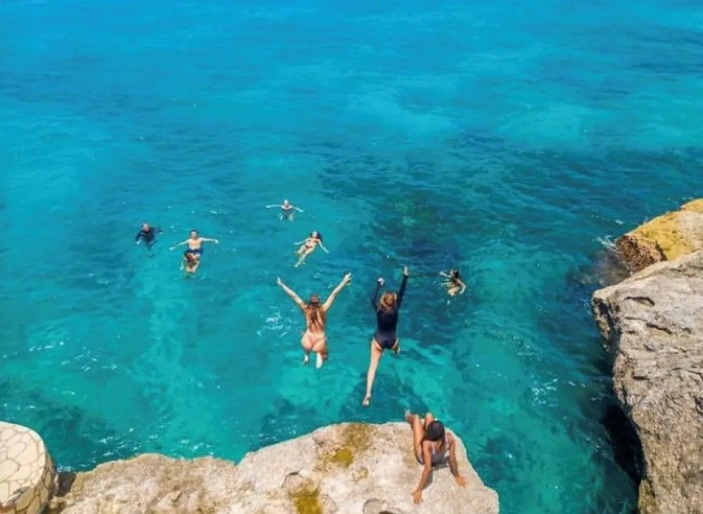 People jump into a clear turquoise ocean from a rocky cliff, arms raised as they swim and splash.