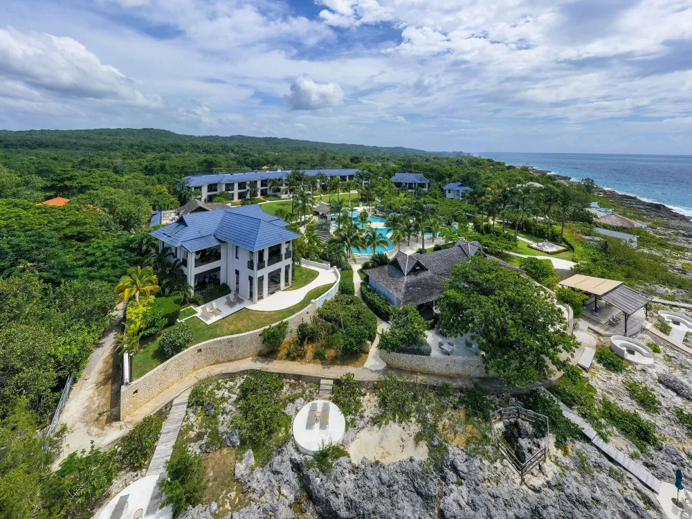 A scenic cliffside overlooking a blue sea with a small white building perched on top, rugged rocks, and green vegetation, under a bright sky.