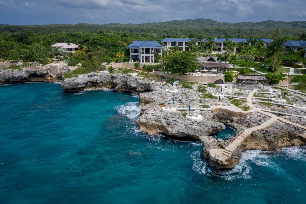 A rocky coastline with clear blue water, small coastal buildings, and a bright sunny sky, overlooking a calm cove.