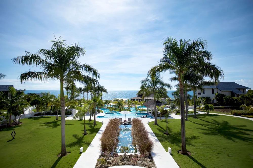 A sunny resort garden with a flagstone path leading to a grand entrance, surrounded by lush green lawn and palm trees.