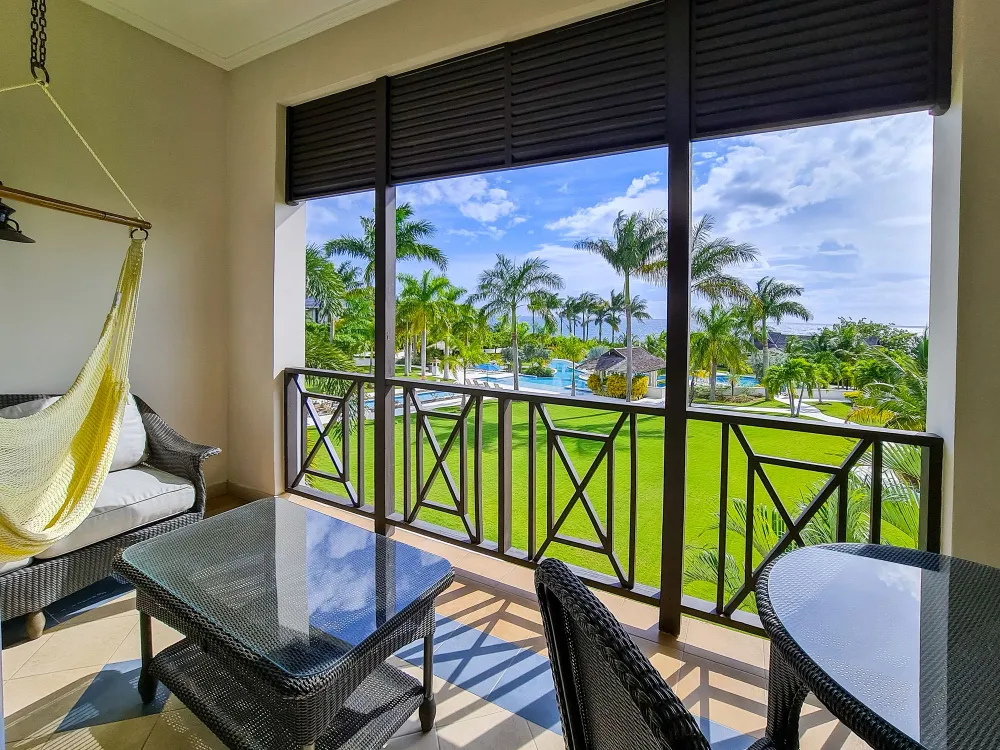 A sunny resort balcony with cushioned chairs, green lawn, palm trees, a pool, and rolling black shutters.