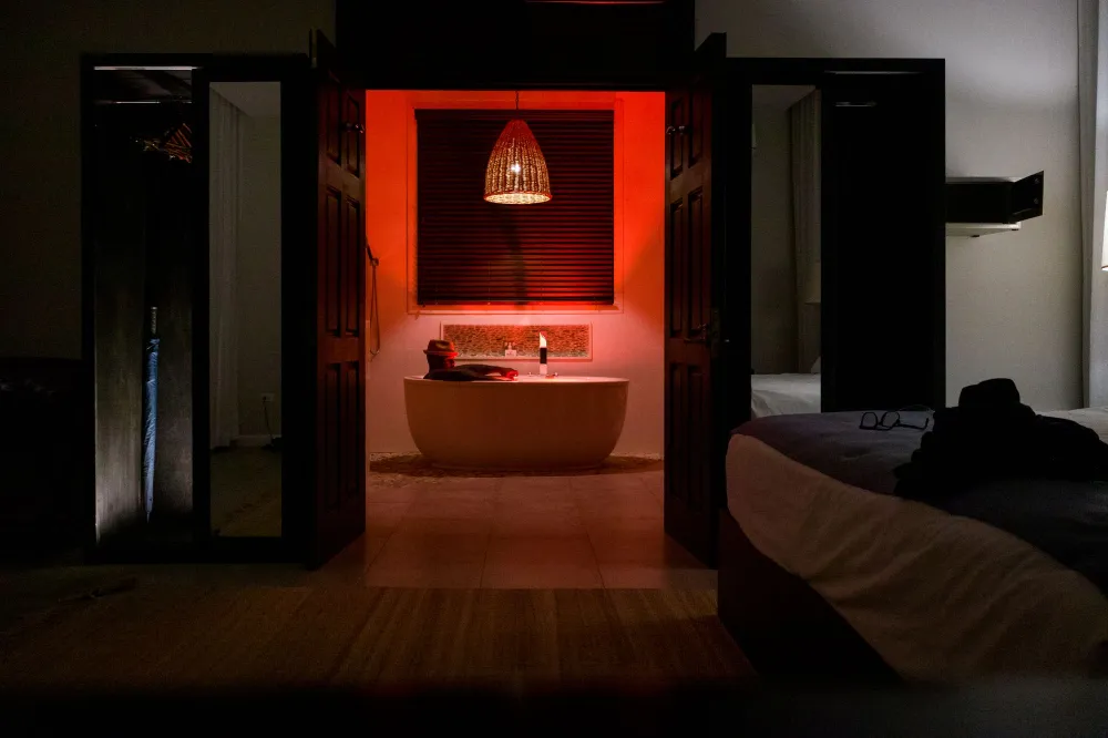 Dim luxury bathroom: red-lit bathtub under a glowing pendant lamp, blind-covered window, and an open doorway to a bedroom.