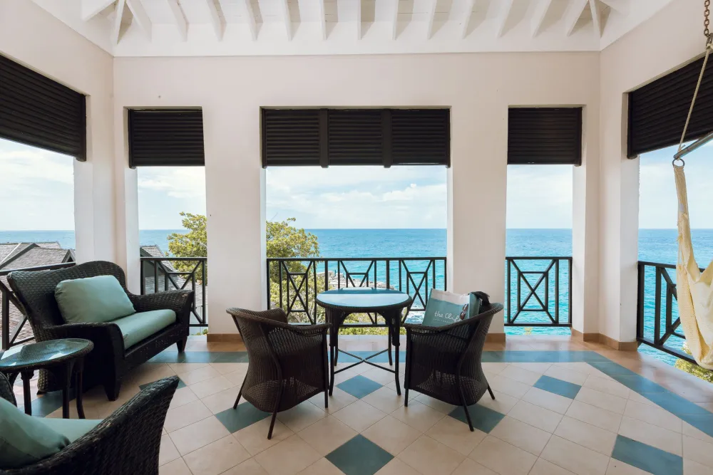 A seaside patio with wicker chairs and table, ocean view through open shutters under a white pergola, with a hanging hammock.