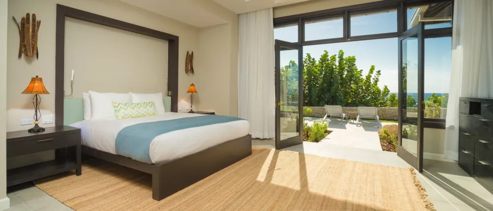 Bright modern bedroom with a bed, lamps, and large sliding doors opening to a patio with chairs and greenery.