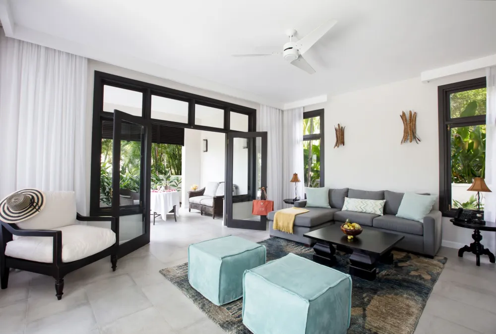 A bright, modern living room with large windows, a plaid sofa, glass coffee table, and a dining area in the background.