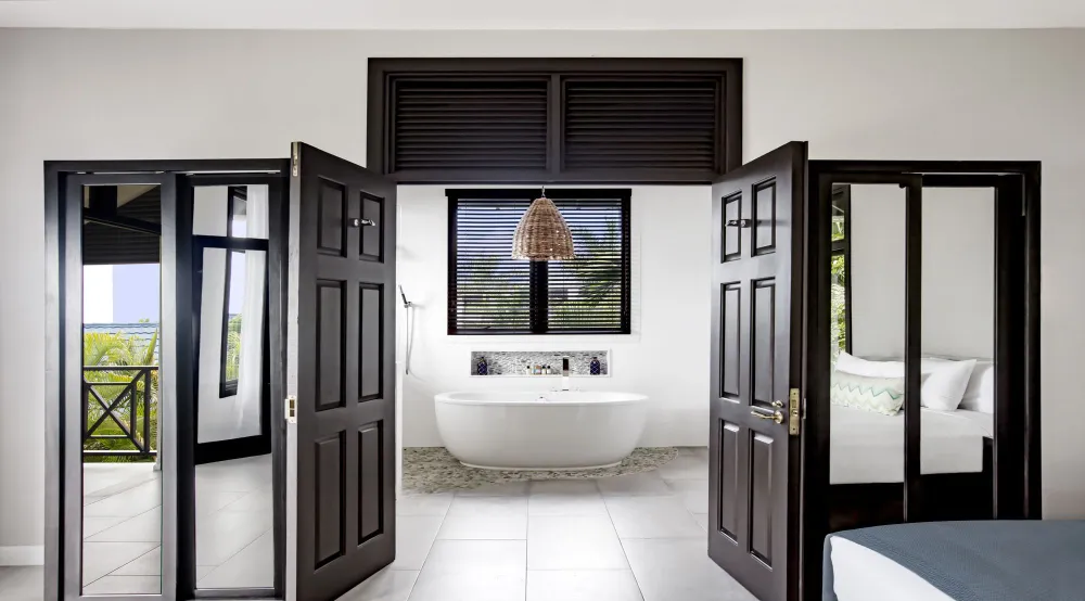 A bright modern bathroom with an open door, oval tub, window blinds, hanging woven light, and a glimpse of the bedroom.