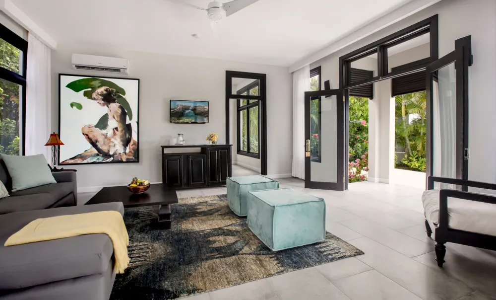 A bright, modern living room with a gray sofa, blue chairs, a white coffee table, and large windows letting in natural light.