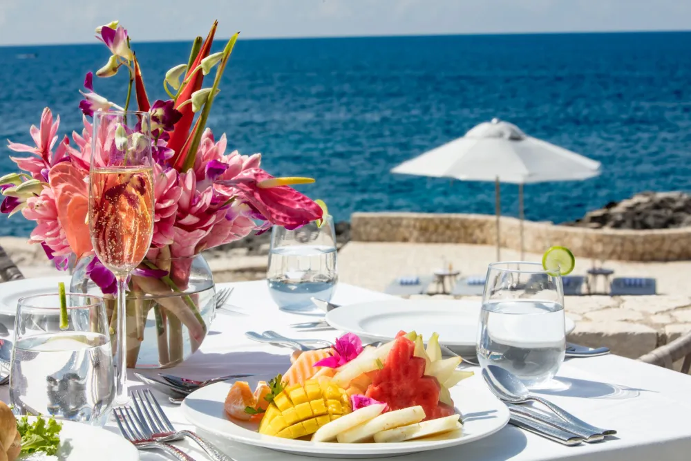 A seaside table set for a meal with tropical flowers, a fruit plate, water glasses, utensils, and a white umbrella over a sunny beach.