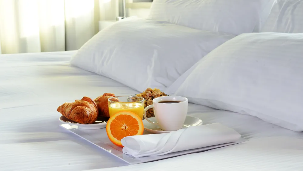 A cozy hotel breakfast on a neatly made bed: croissants, orange juice, coffee, a placed napkin, and a small tray beside fluffy white pillows.
